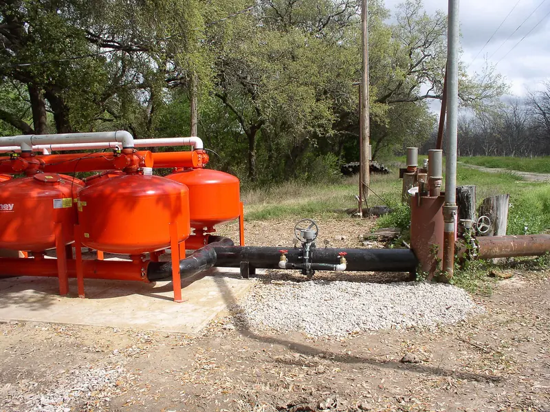 Red filtration vessels and black valved pipework at an irrigation water system among trees on a Texas farm.